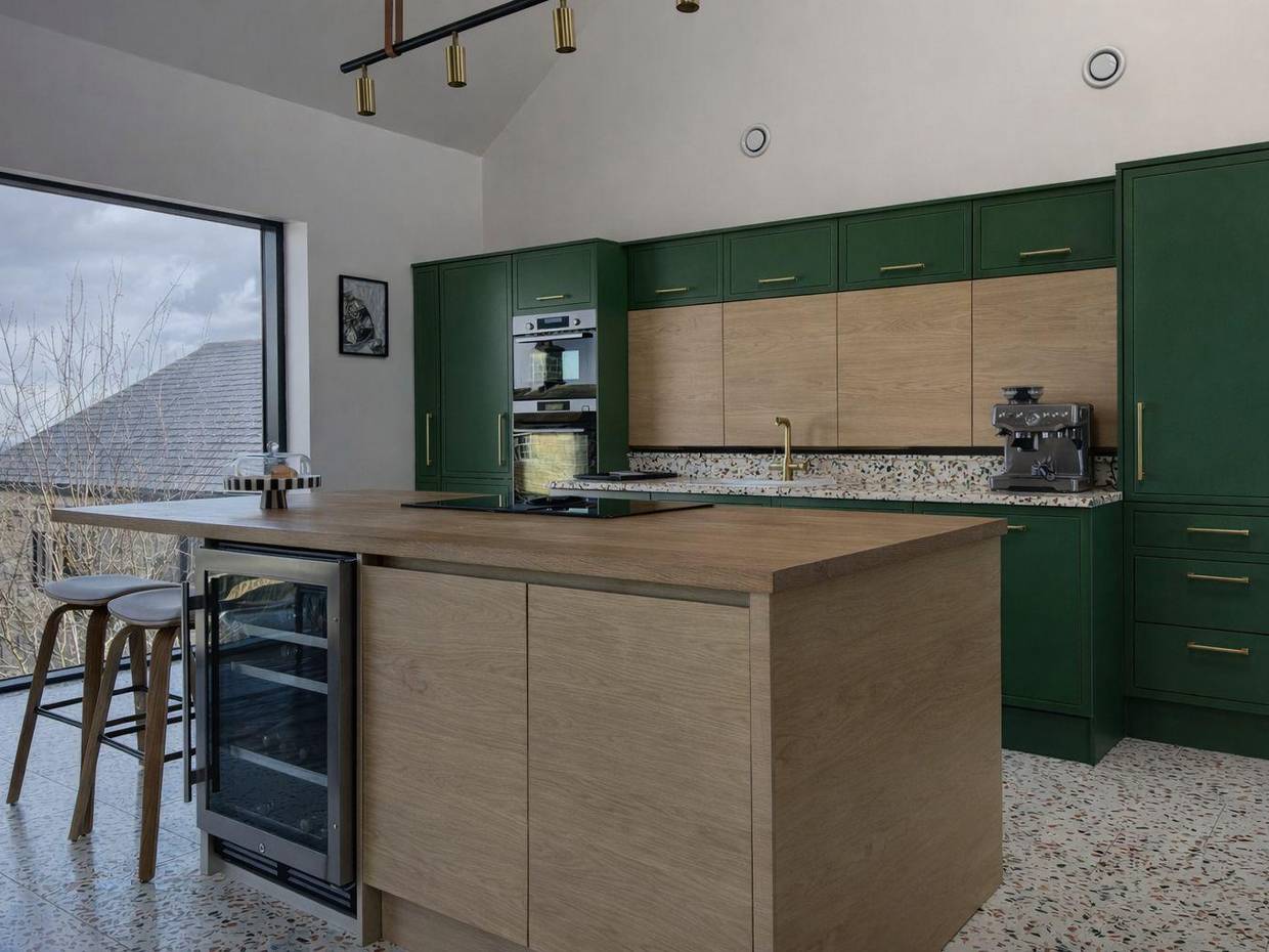 Open-plan kitchen before respraying, with deep forest green units and a wood-fronted island, on a terrazzo floor