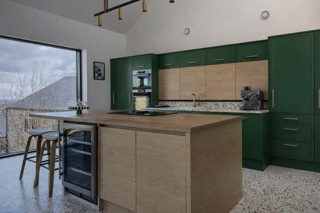 Open-plan kitchen before respraying, with deep forest green units and a wood-fronted island, on a terrazzo floor