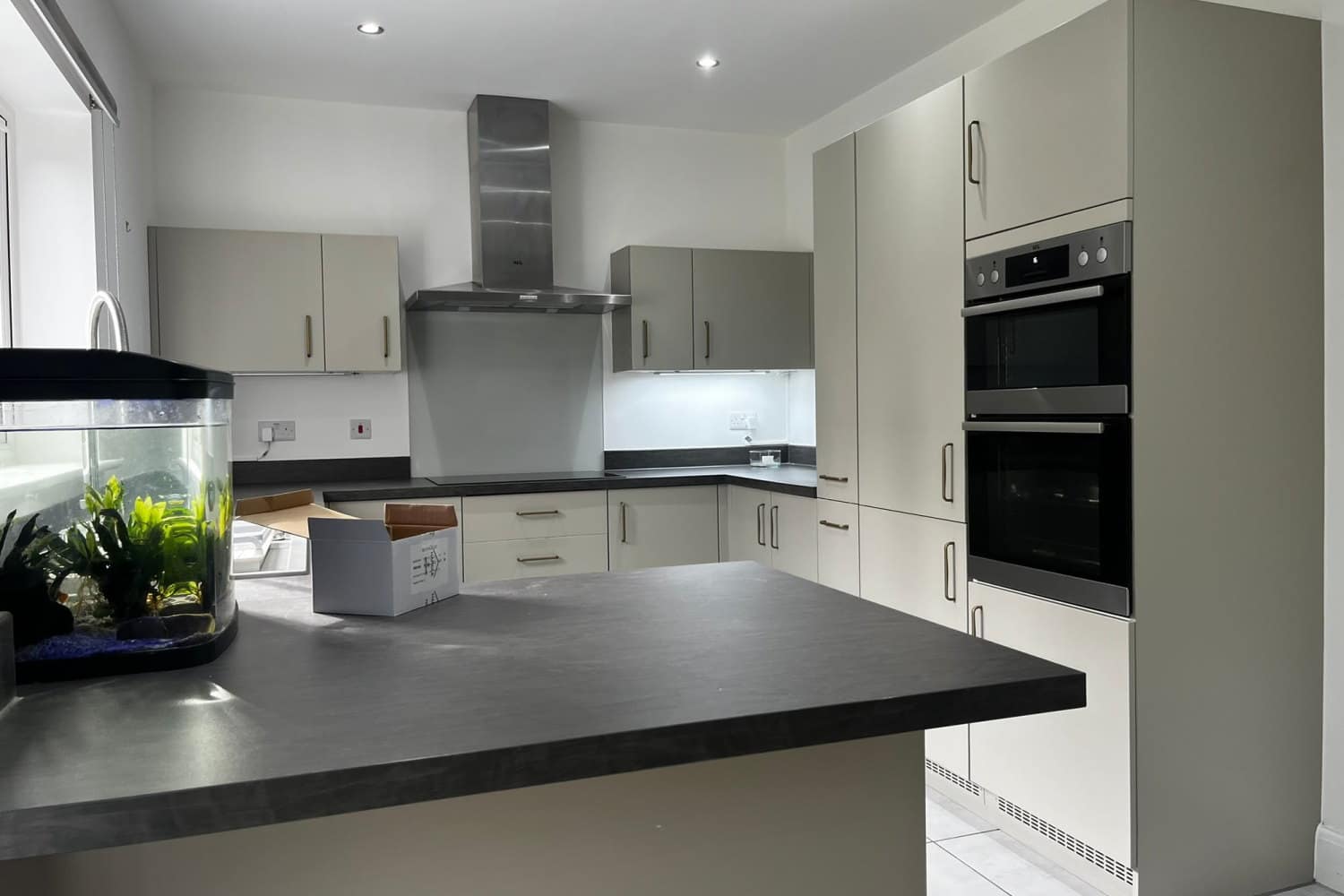A handleless kitchen with an island before respraying, in a pale greige, with a stainless extractor and twin ovens