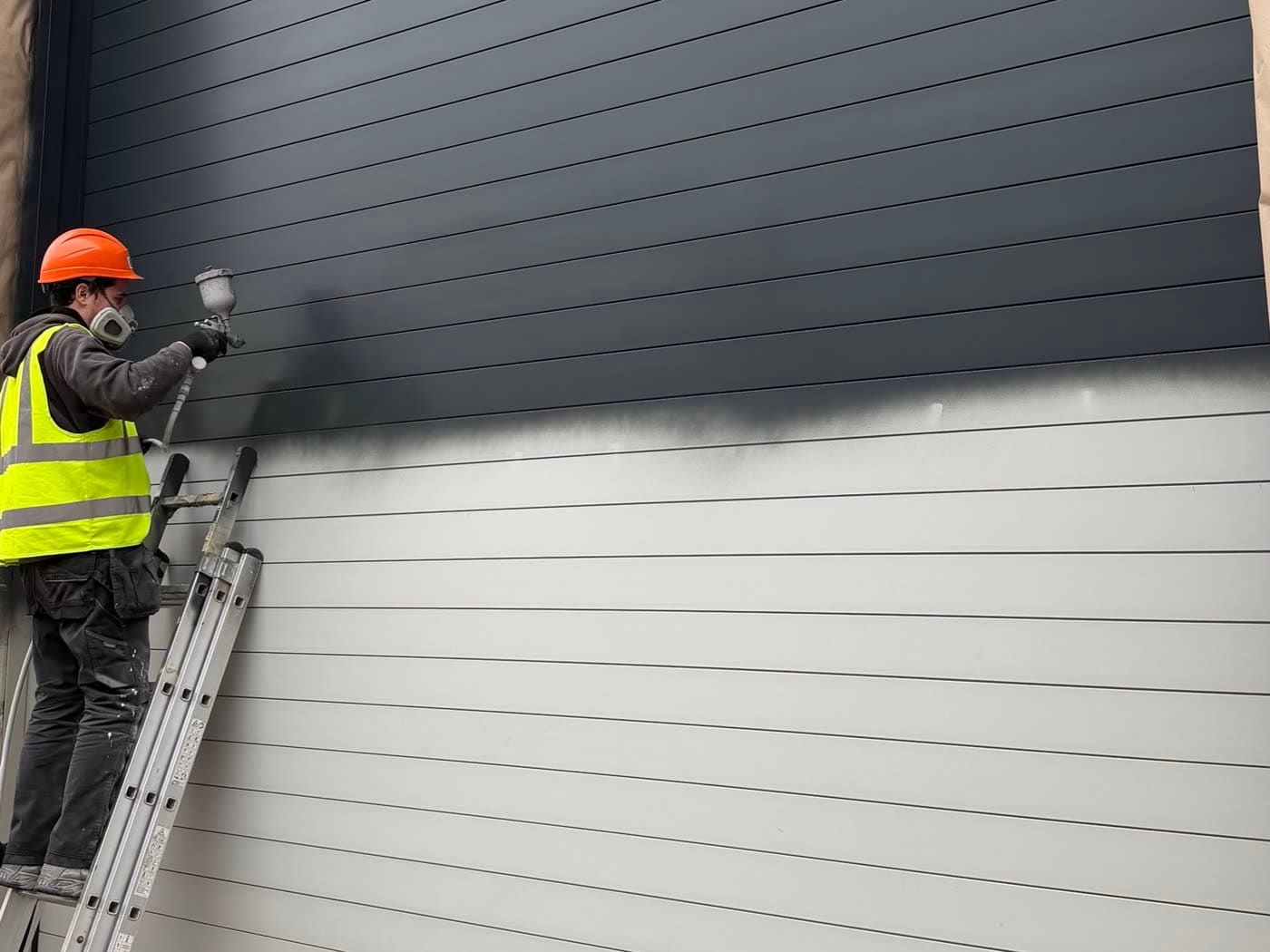 A sprayer on a ladder spraying an industrial roller shutter door in anthracite, the wet edge partway across the white door