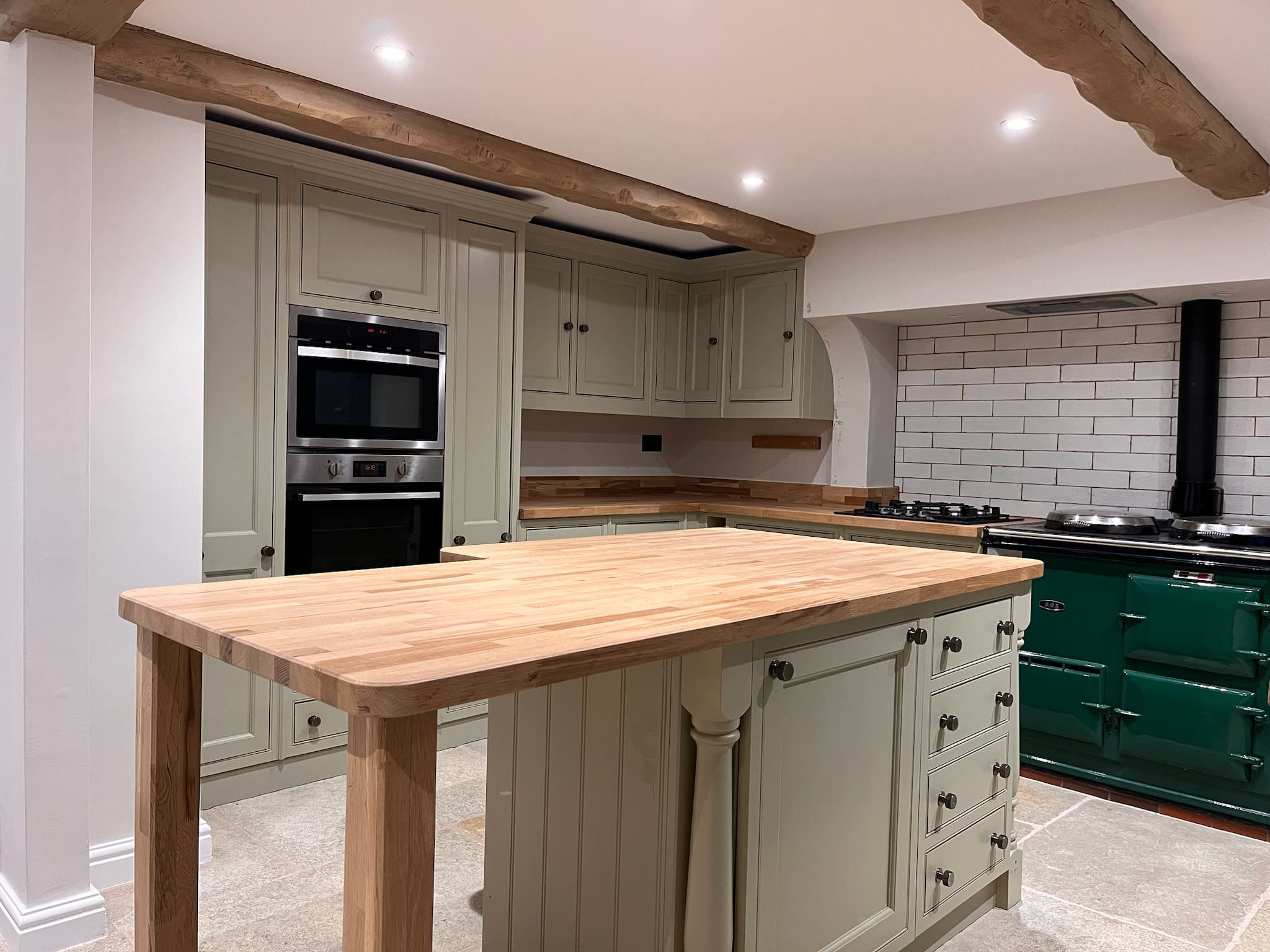 Sage green shaker kitchen respray with an oak beam ceiling, butcher block island and dark green Aga