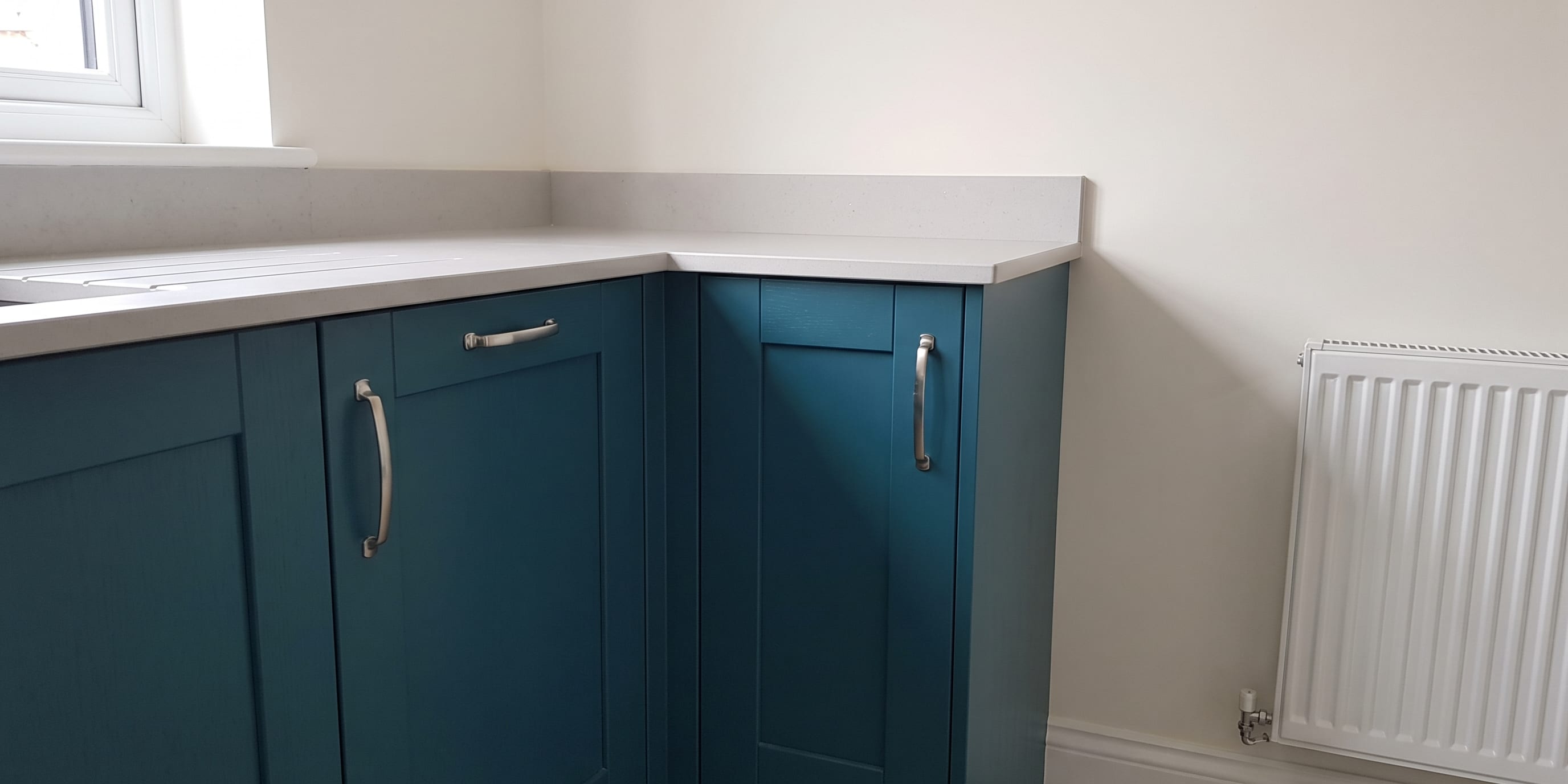 A corner view of teal blue kitchen cabinets with silver handles under a light grey countertop, with a window to the left and a white panel radiator on the wall to the right.