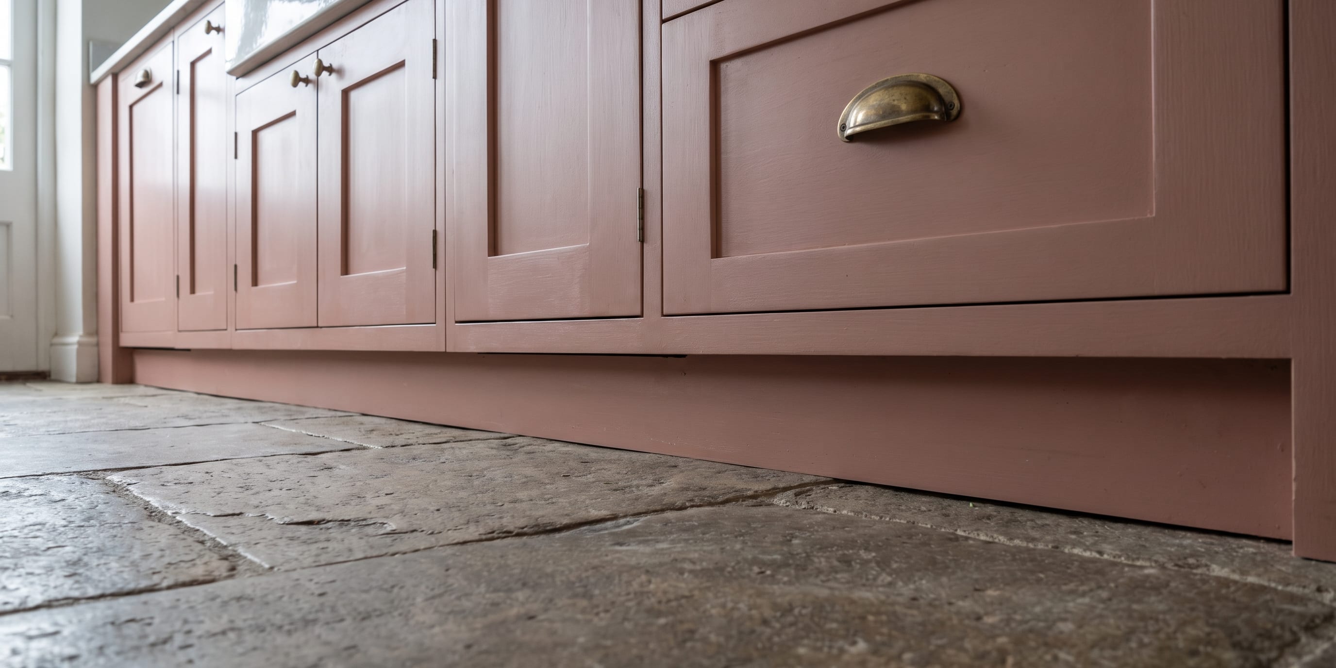 Low-angle view of pink painted shaker-style kitchen cabinets with brass cup handles, standing on a stone flagstone floor.