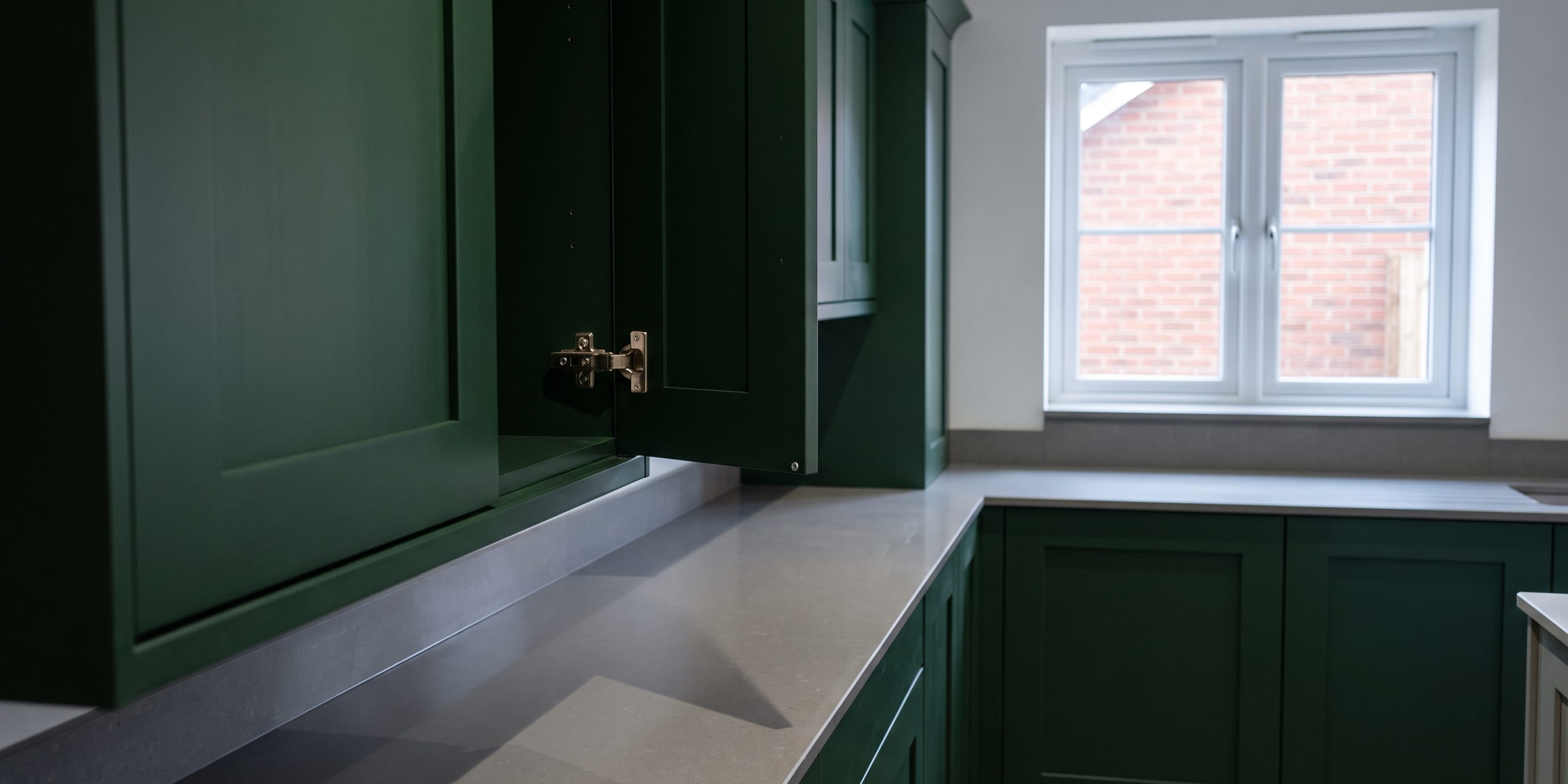 A dark green kitchen with an open upper cabinet door revealing a hinge, a grey countertop, and a window overlooking a brick wall.