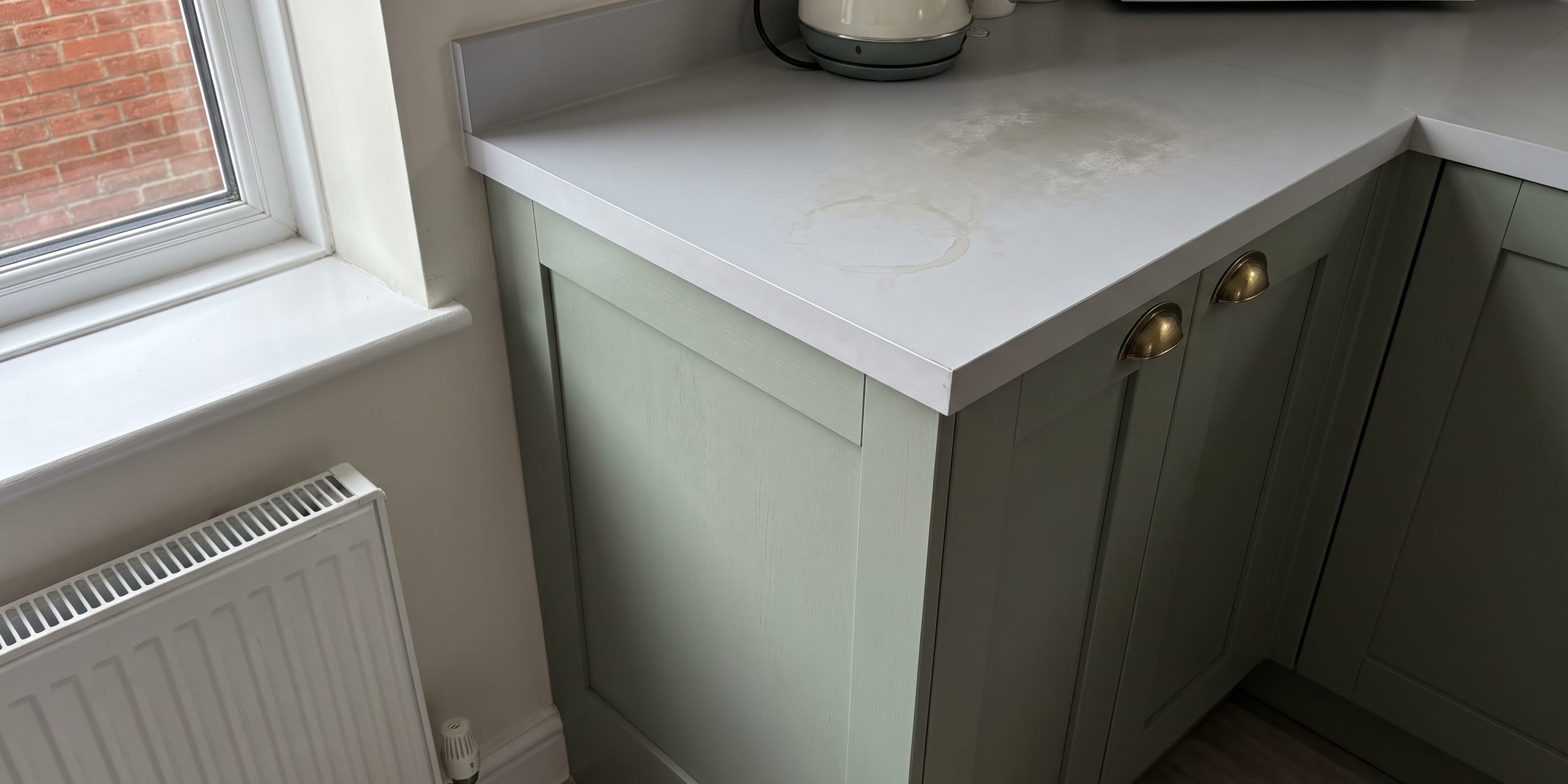 Corner of a white stained kitchen worktop on sage green cabinets with brass cup handles, beside a window and radiator.