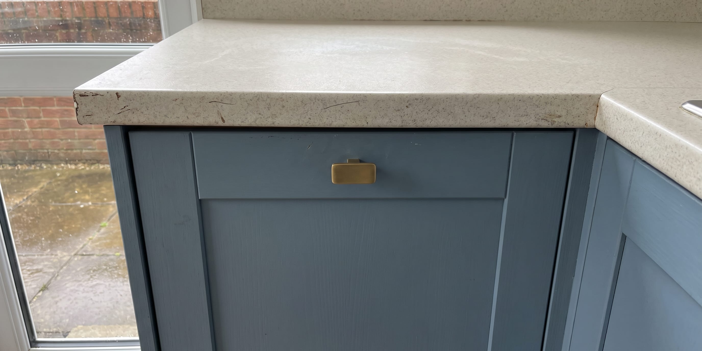 Close-up of a blue shaker-style kitchen cabinet with brass handle beneath a chipped speckled countertop, with a window showing a brick wall outside.