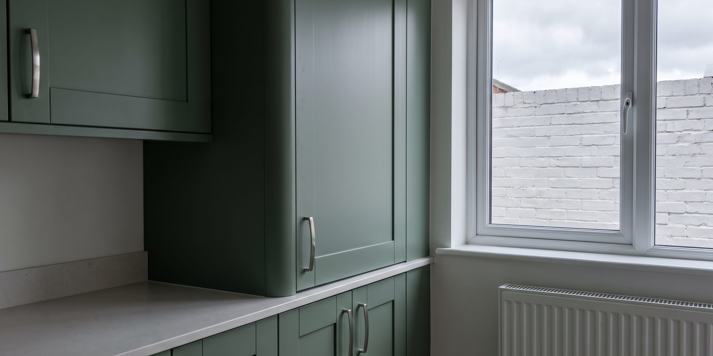 Green shaker-style kitchen cabinets beside a white uPVC window with a wall-mounted radiator below it.