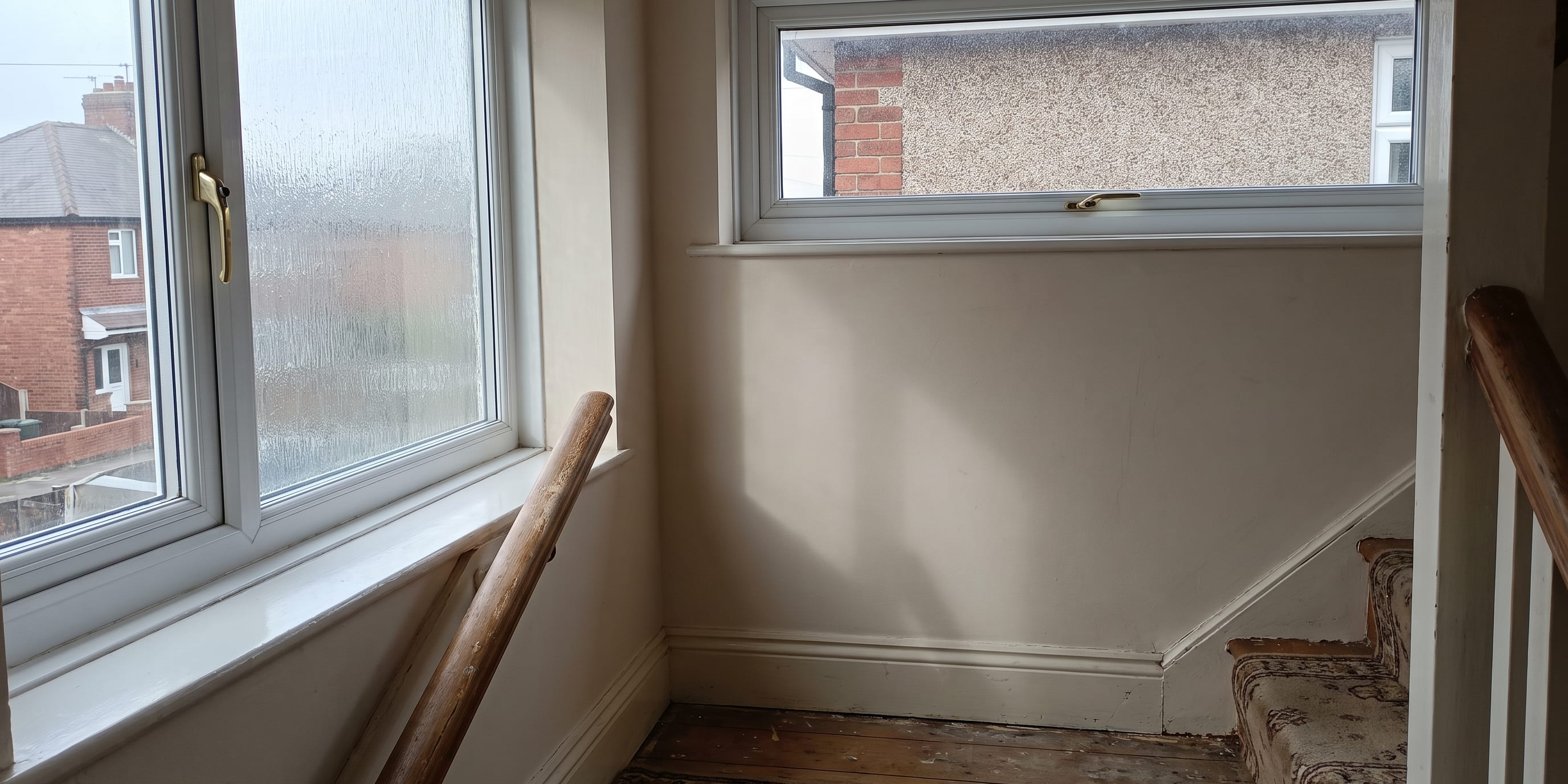 A staircase landing with two white uPVC windows overlooking brick houses, wooden banister and worn carpeted stairs.