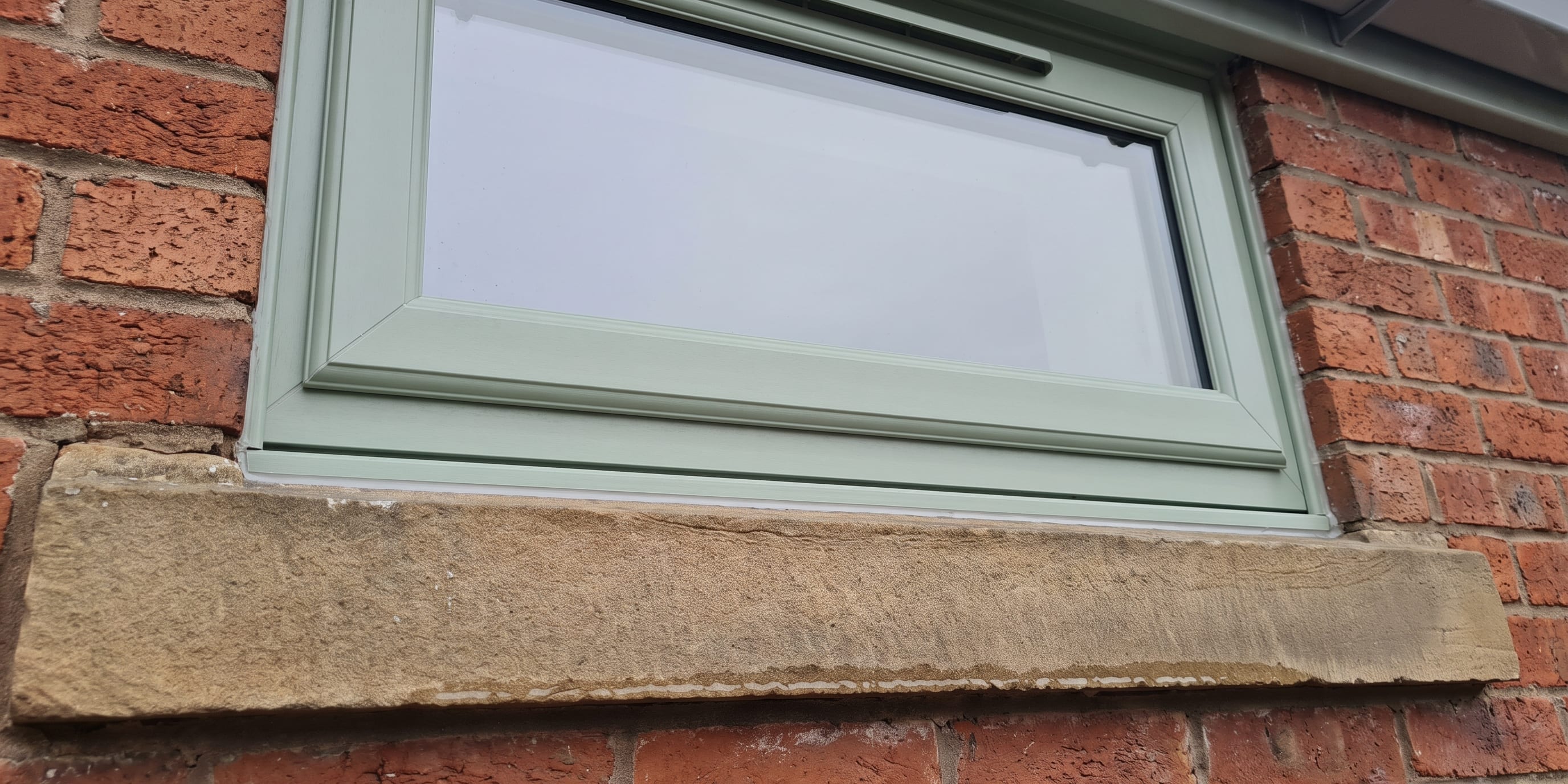 A sage green uPVC window set in a red brick wall with a stone sill below it.